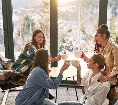 Moment Convivial entre Amies au Chalet de Montagne Quatre amies sourient et trinquent avec des tasses de café dans un chalet de montagne enneigé.