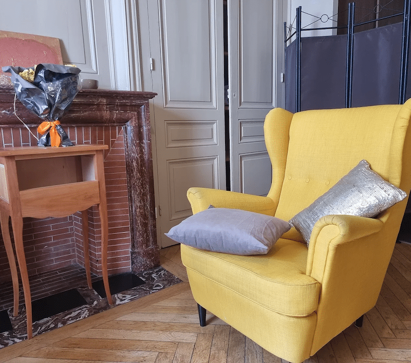 Fauteuil jaune à oreilles et décoration de salon classique Fauteuil jaune à oreilles avec coussins déco devant une cheminée classique et une table de chevet ancienne.