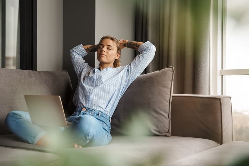 Relaxation et télétravail : femme sereine sur son canapé Femme tatouée se relaxant sur son canapé avec un ordinateur portable, profitant d'un moment de sérénité.