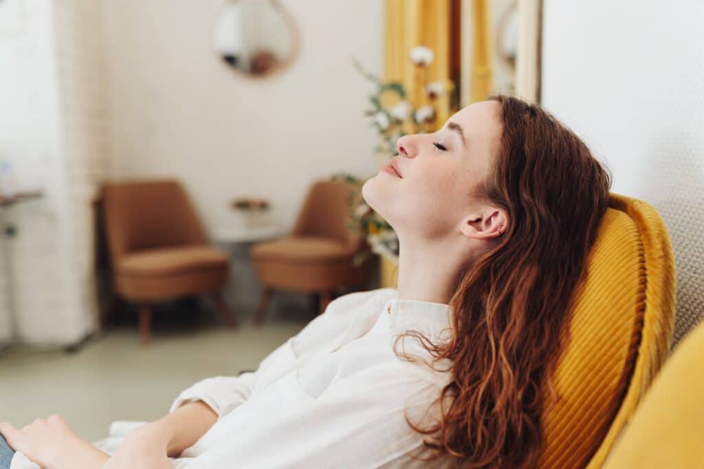Moment de Détente : Femme Rousse Relaxée en Fauteuil Jaune Femme sereine aux cheveux roux se détend dans un fauteuil jaune en velours, yeux fermés, moment de bien-être.