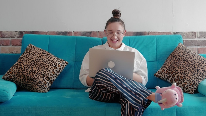 Télétravail à domicile : femme souriante sur canapé bleu Femme souriante avec lunettes utilisant un ordinateur sur un canapé bleu, chaussons licorne et coussins léopard.
