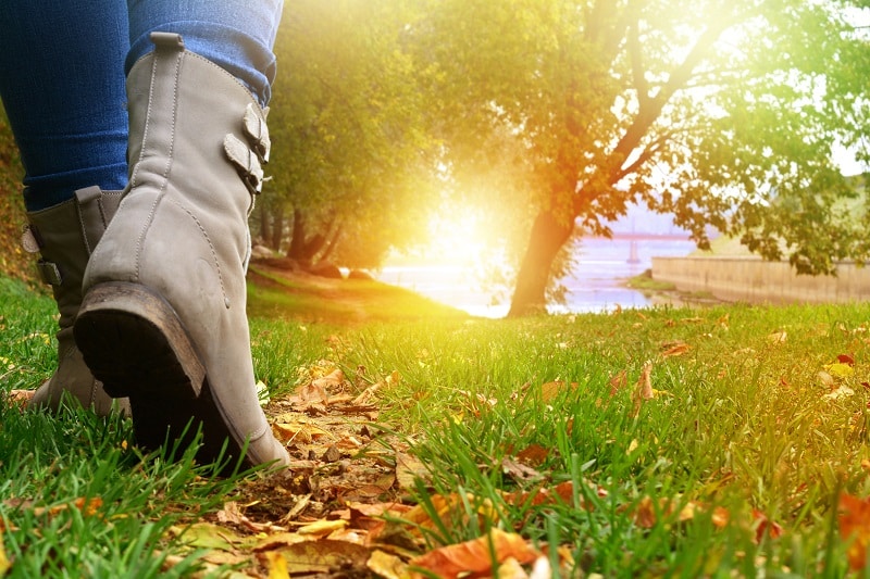 Marche Automnale en Bottines dans un Parc au Soleil Une personne marche en bottines grises sur l'herbe et les feuilles d'automne dans un parc ensoleillé au bord de l'eau.