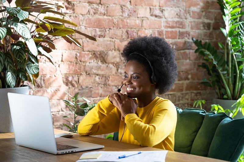 Visioconférence : Femme souriante travaillant en télétravail Femme afro souriante avec casque micro en visioconférence sur ordinateur portable, bureau avec plantes.